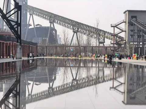 镜面水景倒映出钢铁桁架、厂房与游人
